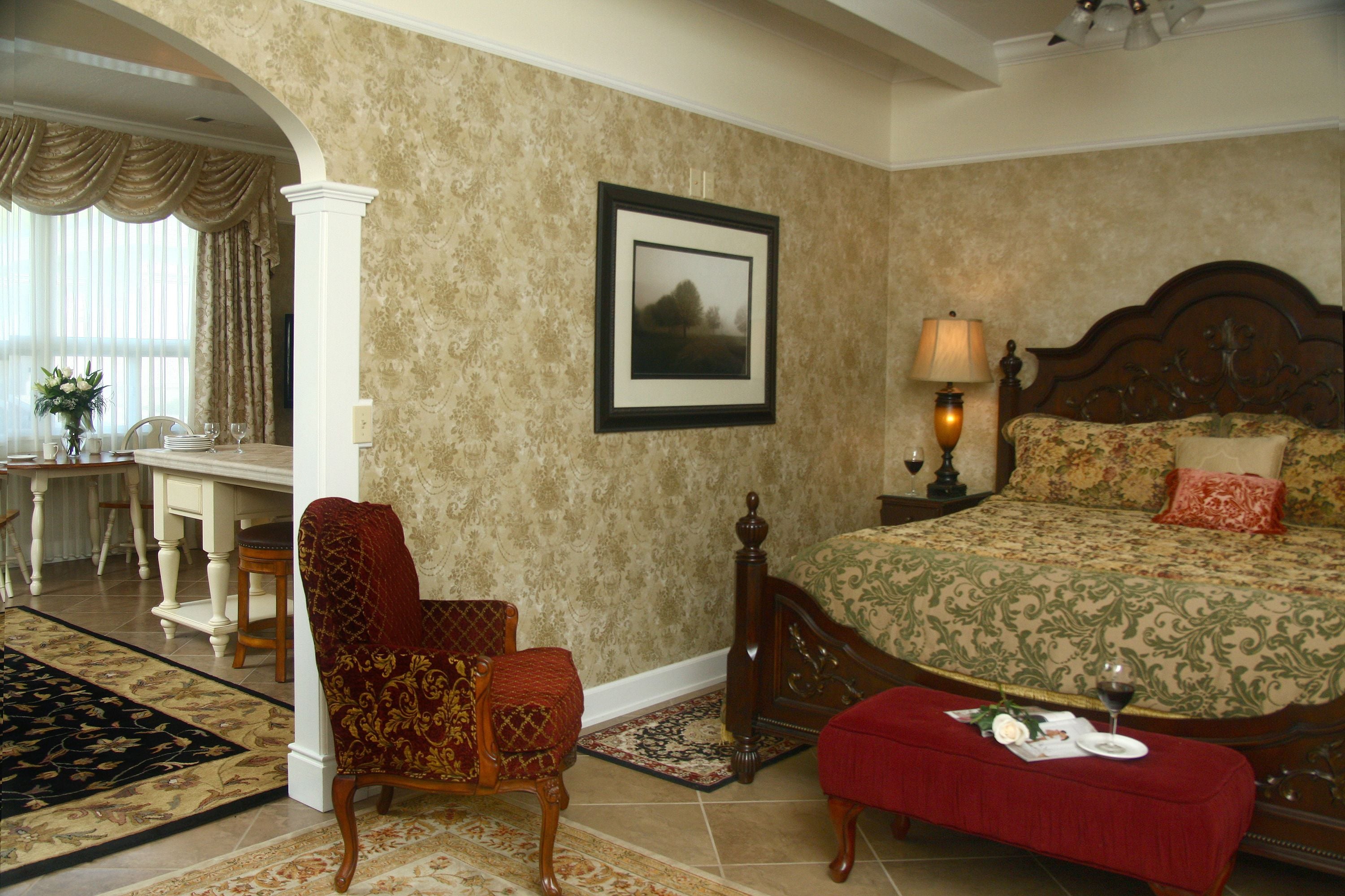 A bedroom with a bed, a chair, a table with a lamp, and a framed picture on the wall.