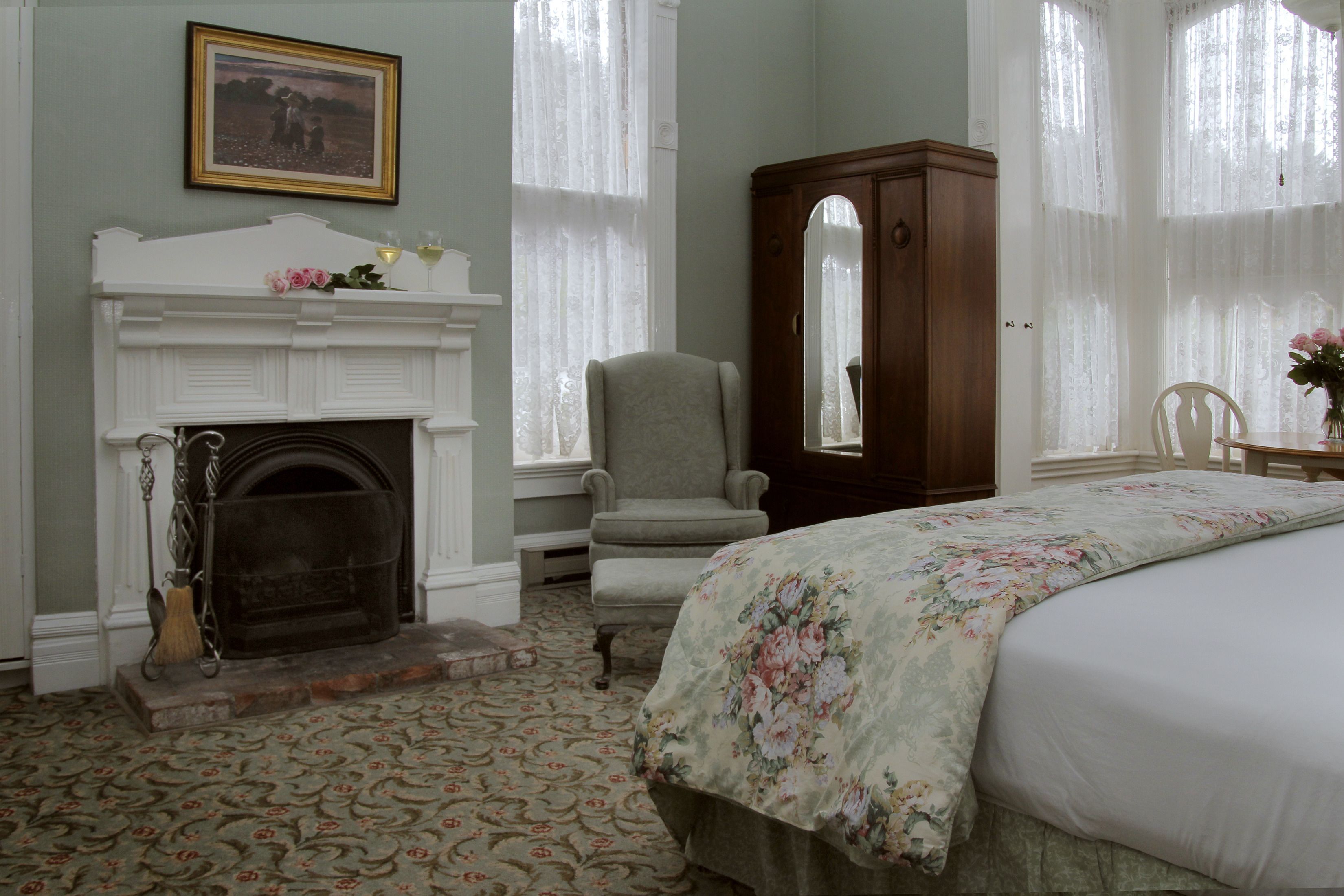 A bedroom with a fireplace, a large bed with a floral bedspread, a wooden armoire, and a chair.