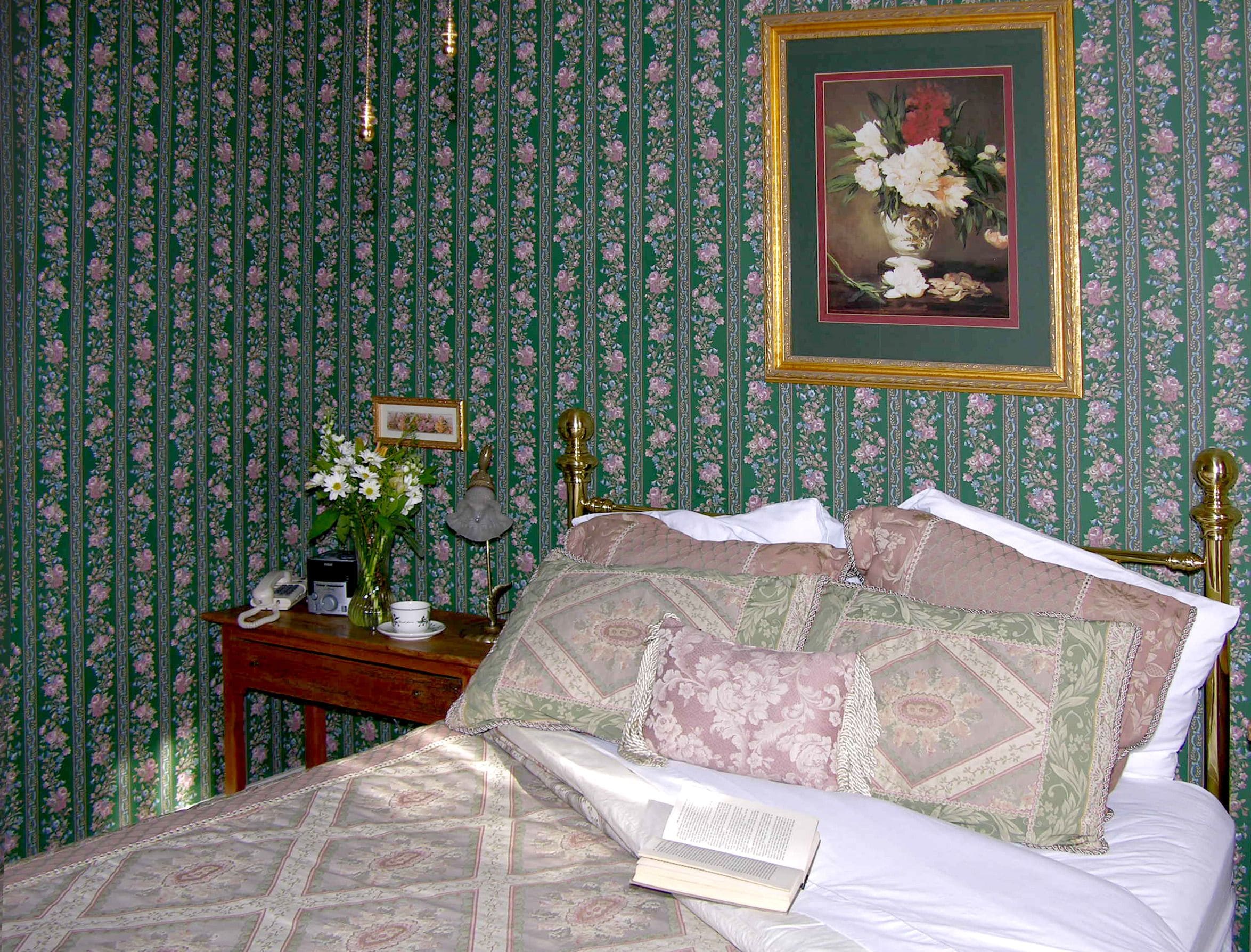 A bedroom with floral wallpaper, a bed with patterned bedding, a bedside table with a lamp, and a framed painting above the bed.
