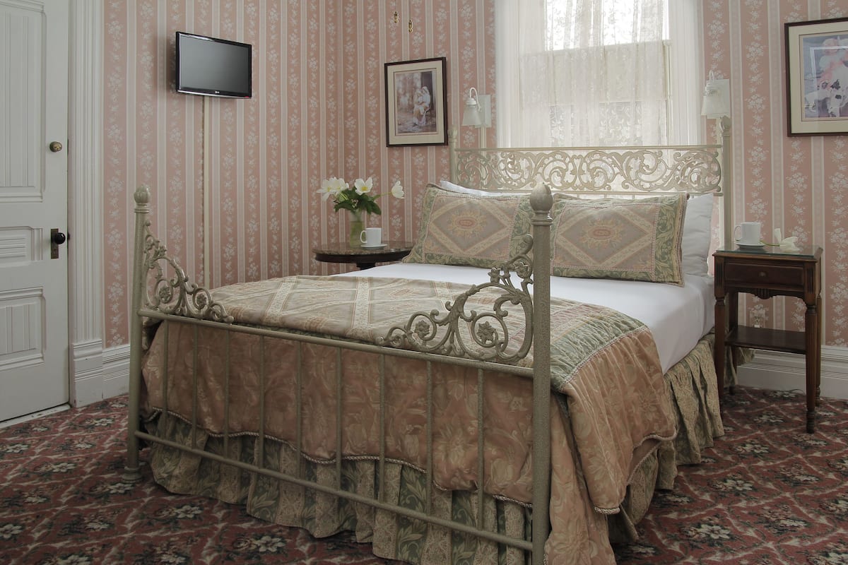 A classic four-poster bed with a floral bedspread, a bedside table with a vase of flowers, and a framed picture on the wall.