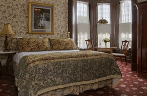 A bedroom with a large bed, a desk with a chair, a lamp, and a painting on the wall.