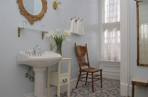 A bathroom with a pedestal sink, a wooden chair, a mirror with an ornate frame, and a window with sheer curtains.