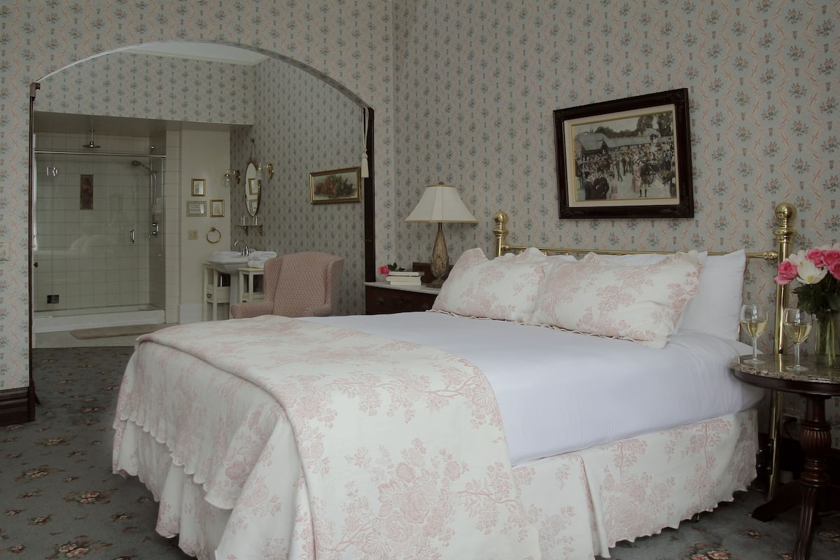 A bedroom with a large bed, a bedside table with a lamp, a mirror, and a picture on the wall. There is a bathroom visible in the background.