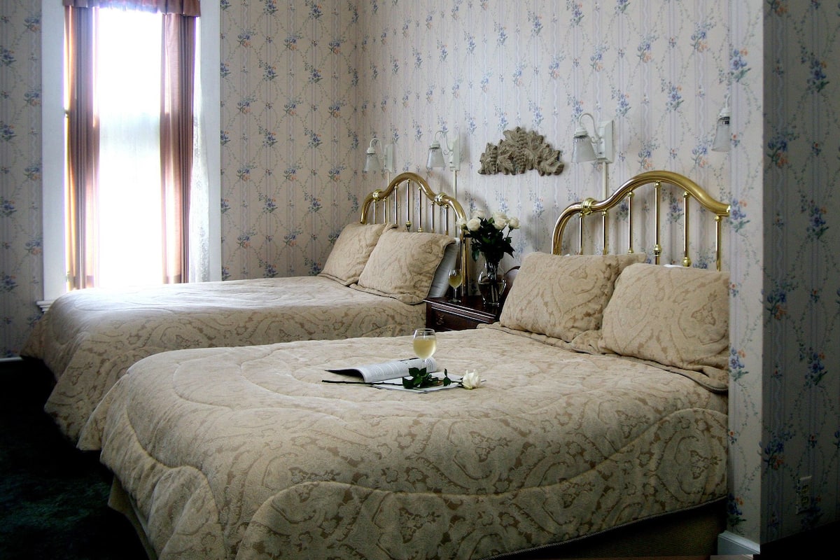 A hotel room with two beds, floral wallpaper, and a window with curtains.