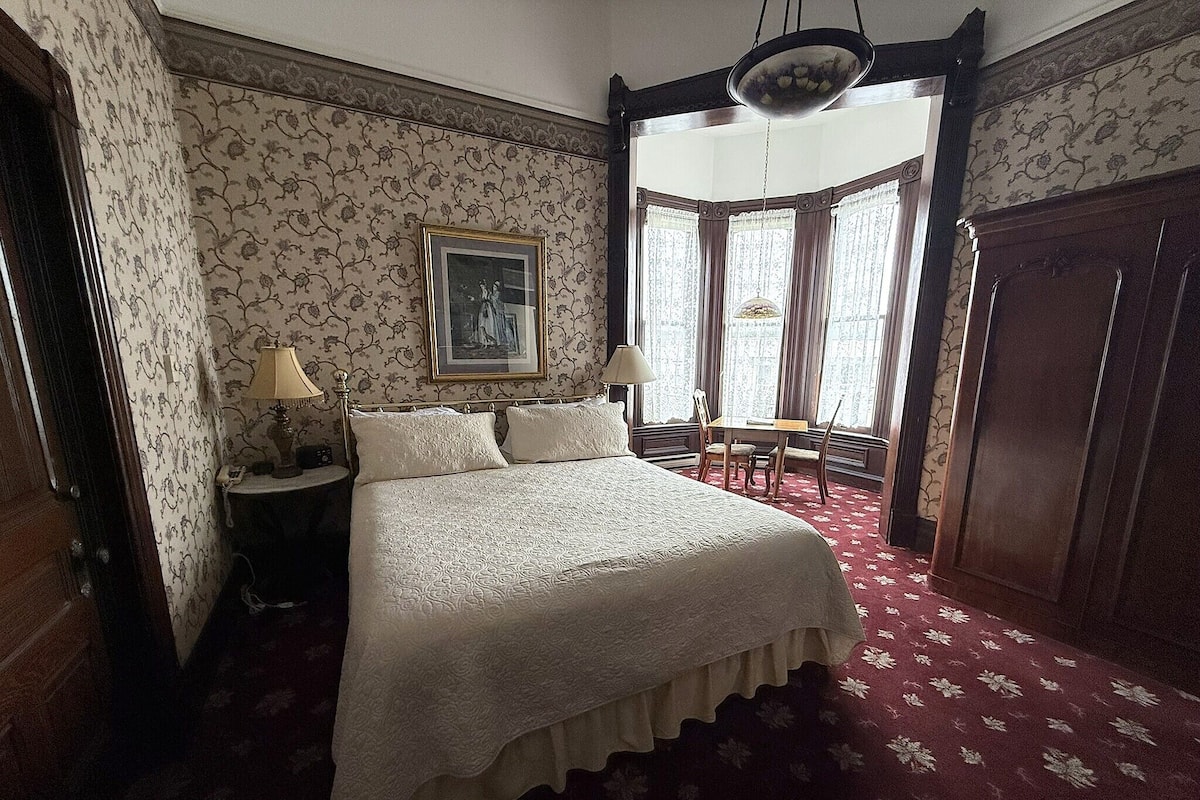 A bedroom with a large bed, a bedside table with a lamp, a framed picture on the wall, and a window with curtains.