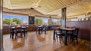 Restaurante com piso, mesas e cadeiras de madeira. Janelas amplas oferecem vista para árvores e montanhas.