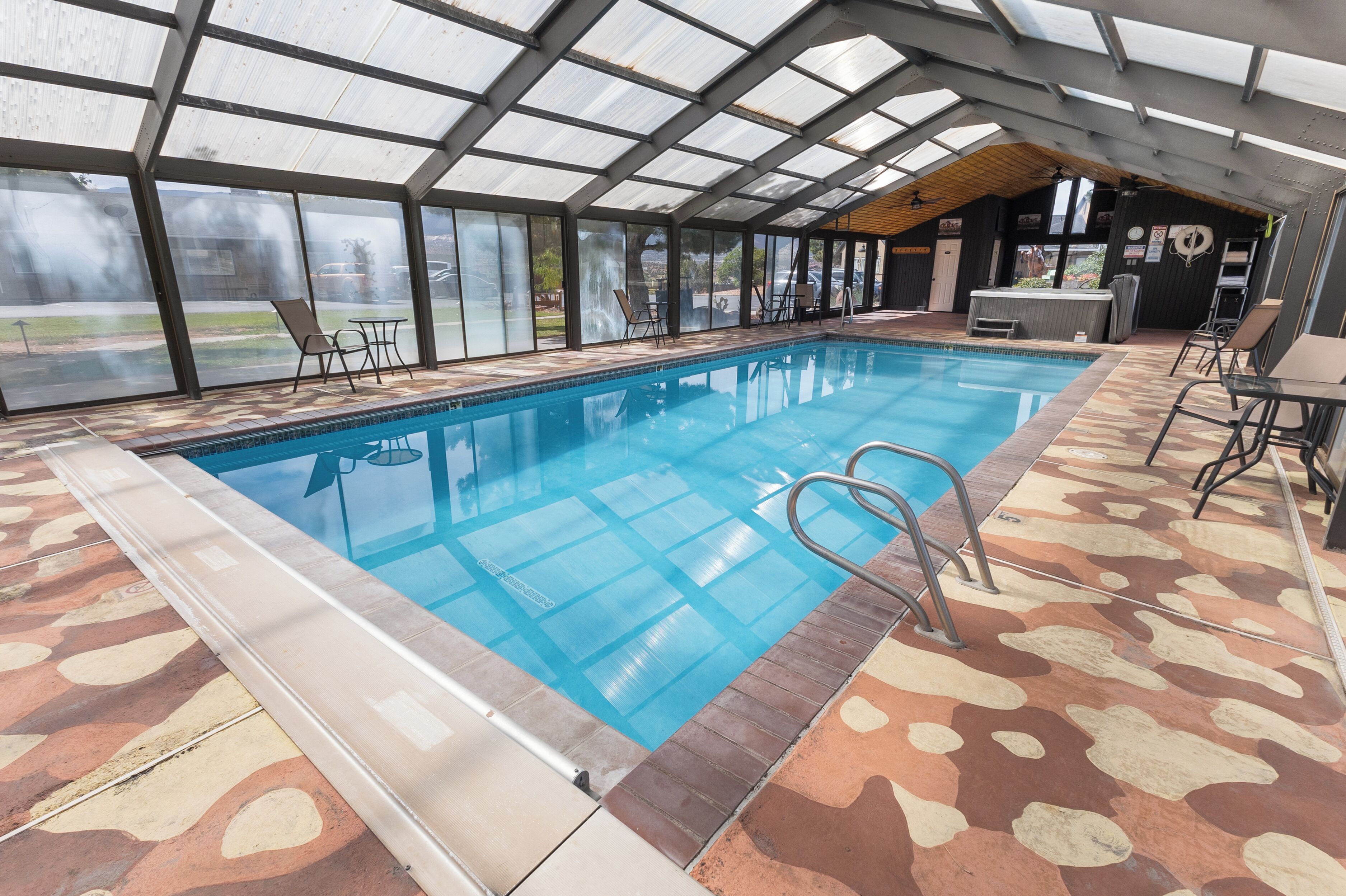 An indoor swimming pool with clear glass walls, a pool ladder, and a patterned floor.