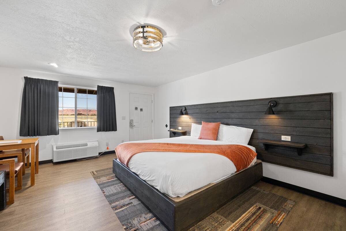 A modern bedroom with a large bed, a wooden headboard, a desk, and a view of the desert.