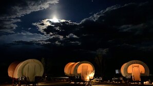 Três tendas em formato de cúpula com uma fogueira central, localizadas em um campo sob um céu noturno com nuvens e uma lua visível.
