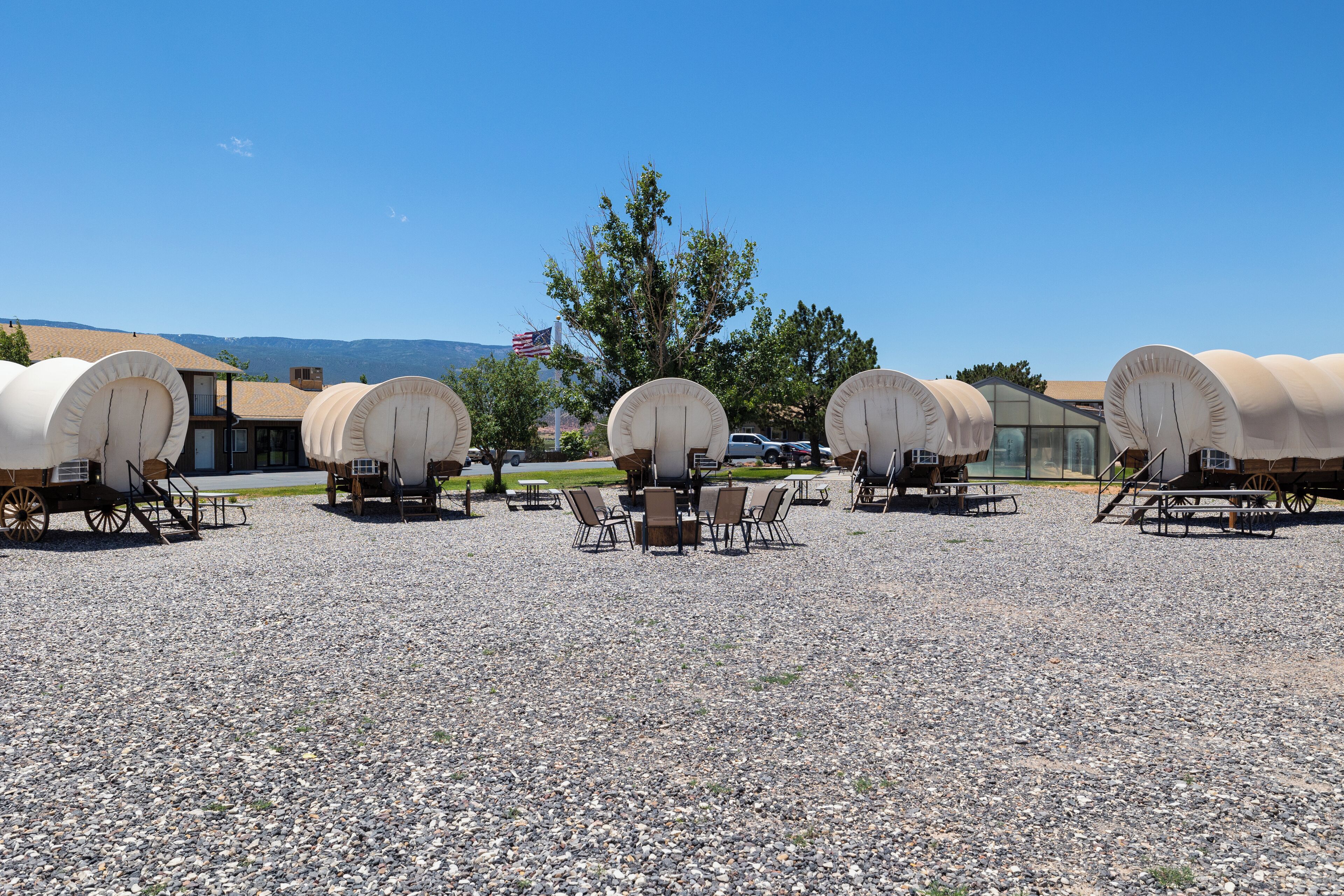 A series of covered wagons arranged in a row with tables and chairs in between, set on a gravel surface with a clear sky and mountains in the background.
