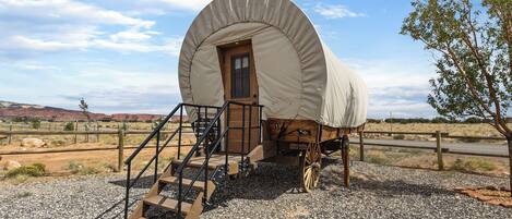 A covered wagon with a wooden door and steps leading up to it, set on a gravel surface with a fence in the background.
