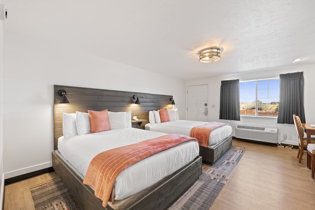 A hotel room with two beds, a wooden headboard, a large window with curtains, a desk, and a chair.