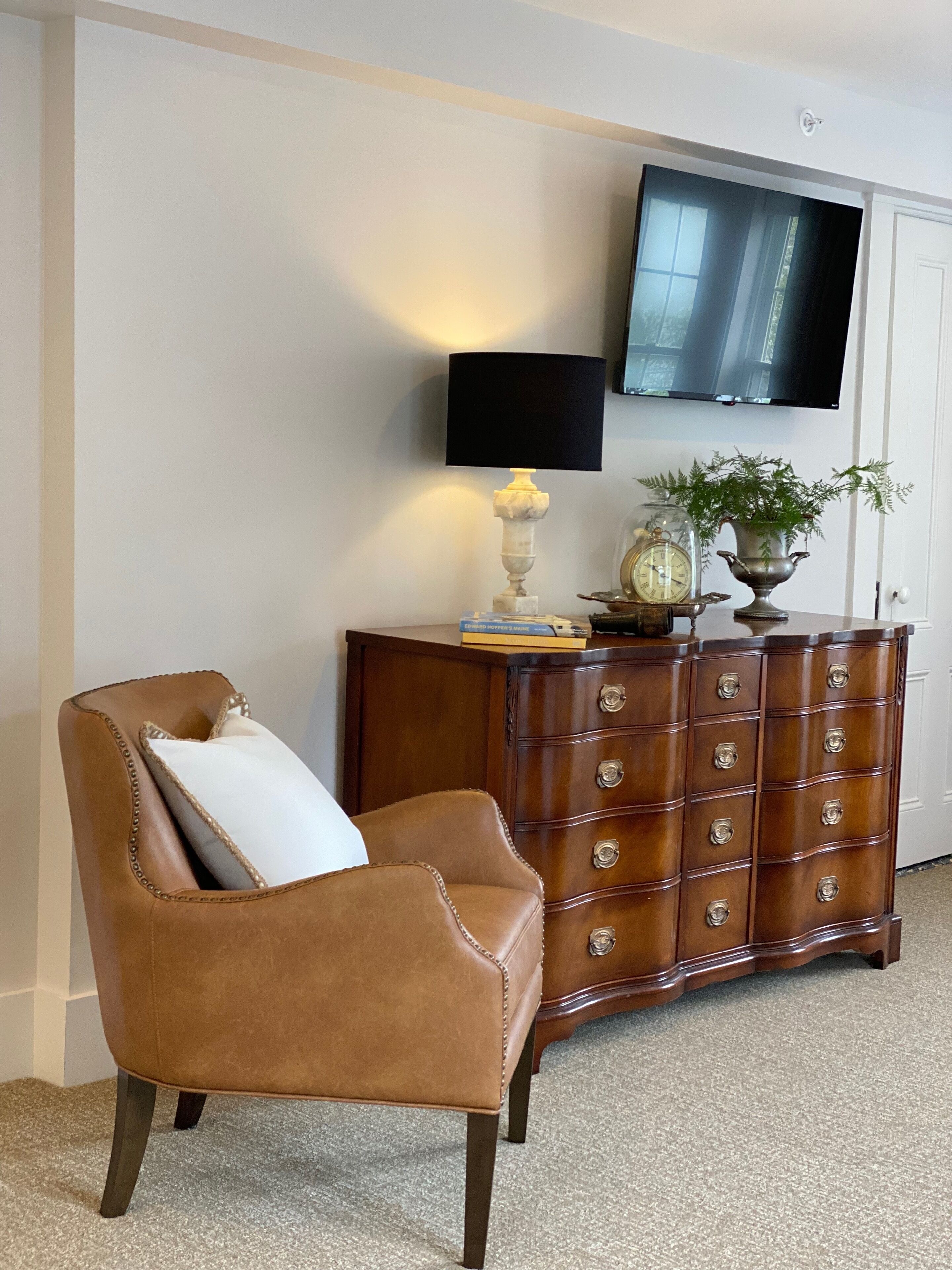 Un sillón de cuero marrón con un cojín blanco, un aparador de madera con varios cajones, un televisor montado en la pared y una planta pequeña sobre el aparador.