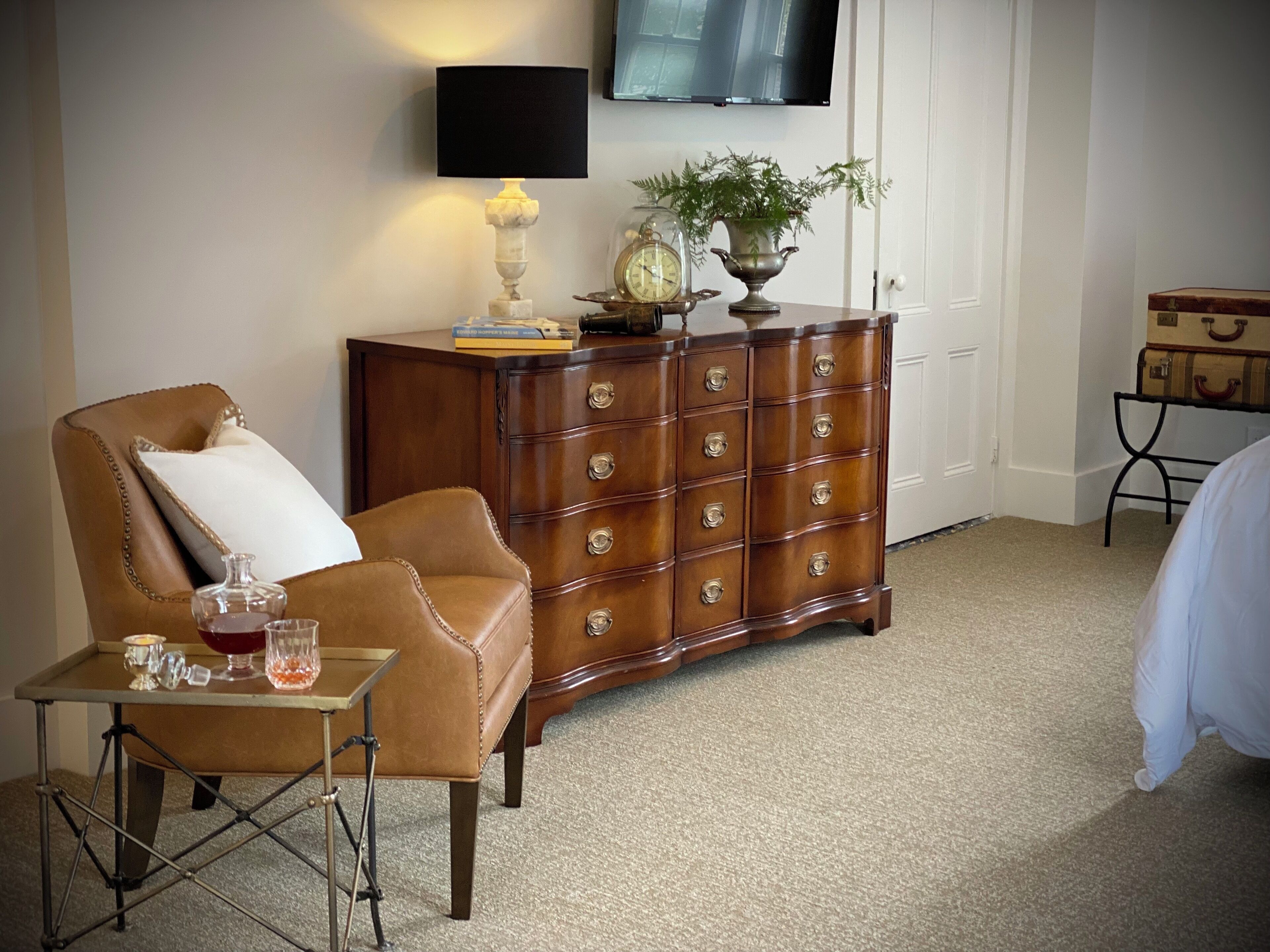 Een kamer met een leren fauteuil, een houten dressoir, een klein tafeltje met een lamp en een aan de muur bevestigde televisie.