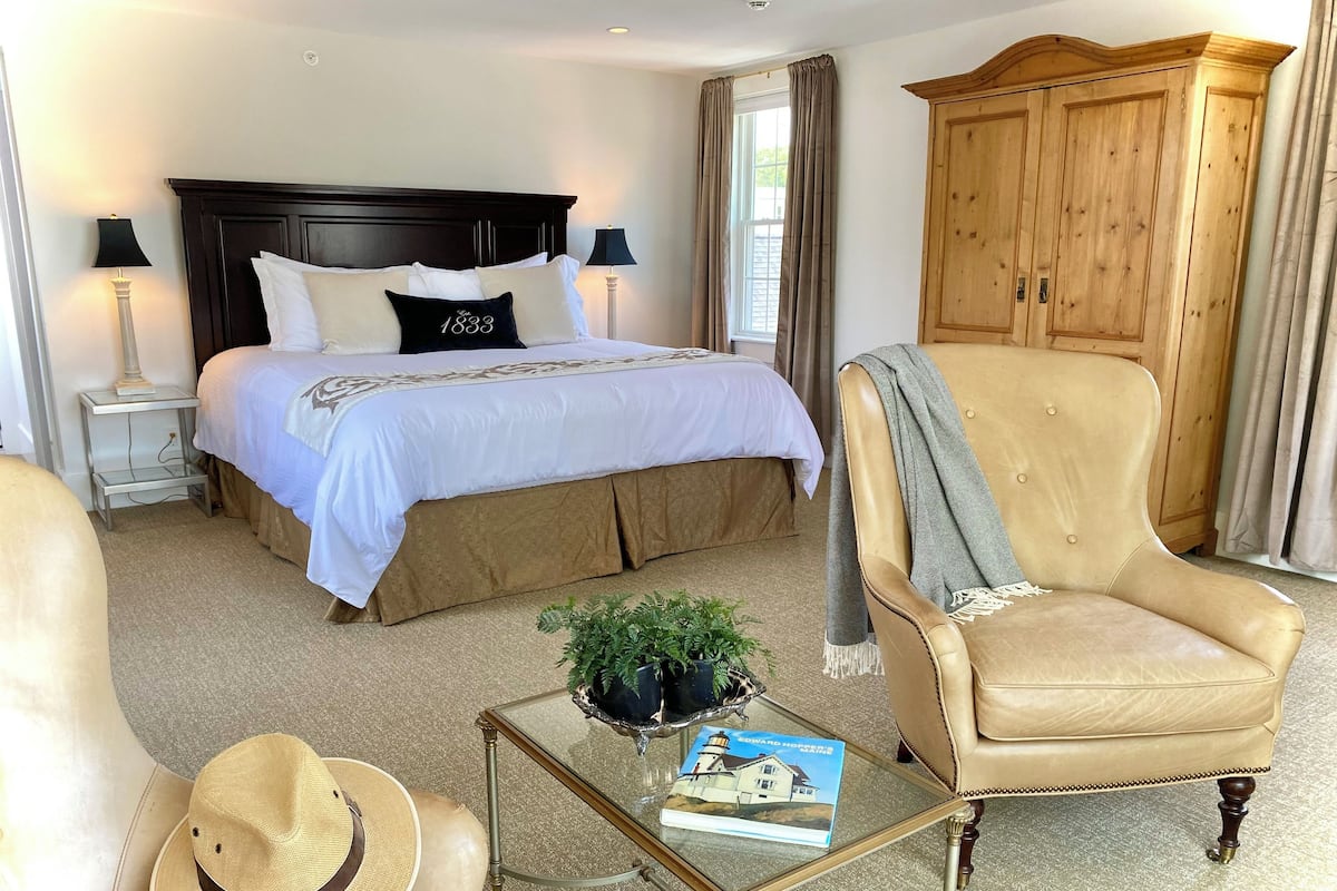 A hotel room with a large bed, a wooden wardrobe, a leather armchair, a glass coffee table with a book and a hat, and a small side table with a lamp.