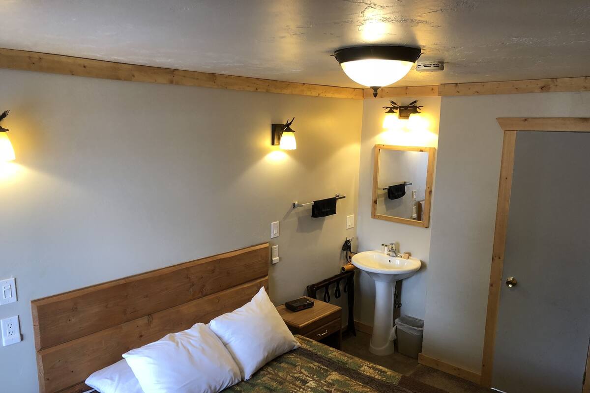 Un dormitorio con cabecera de madera, una cama con almohadas blancas, una mesita de noche y una lámpara, un espejo montado en la pared, un lavabo y una puerta.