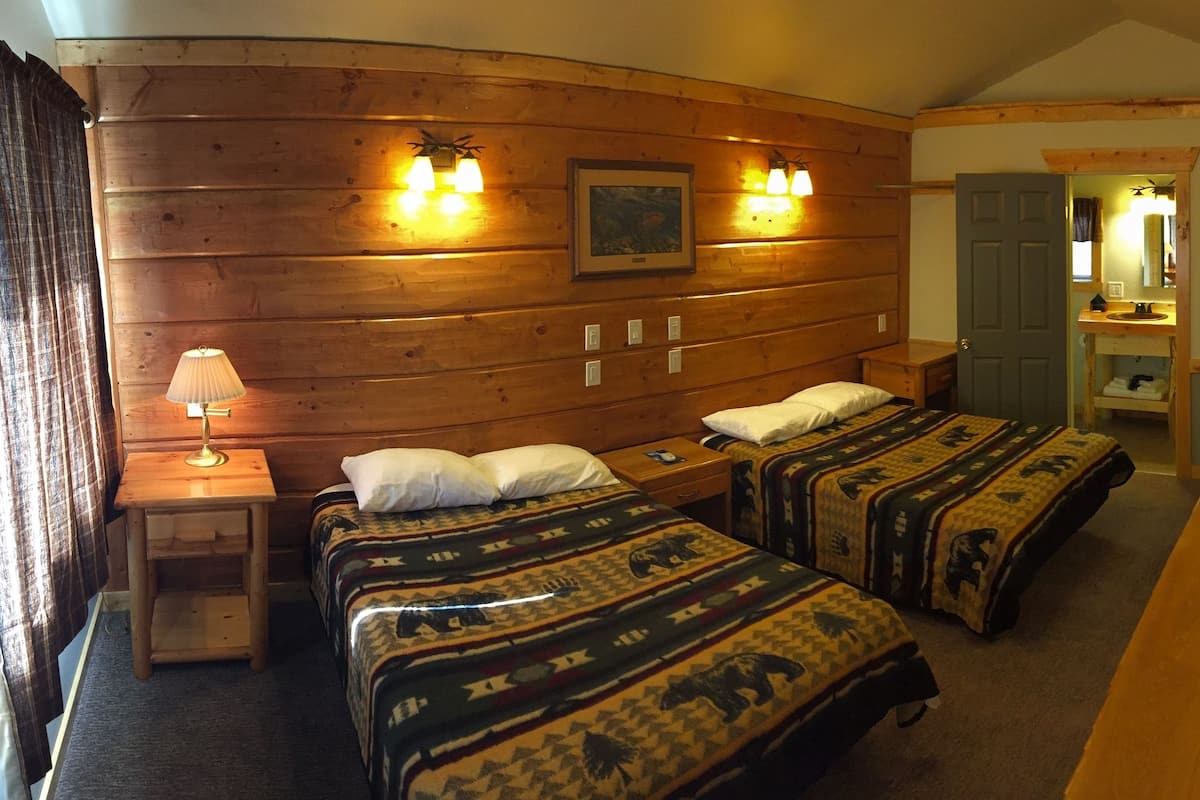 Una habitación tipo cabaña de madera con dos camas, una mesita, una lámpara y un banco de madera.