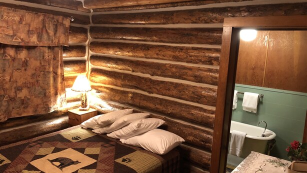 A log cabin room with a bed, a nightstand with a lamp, a quilt, a wooden wall, a bathroom with a sink and a bathtub. - The Historic Madison Hotel Motel (West Yellowstone)