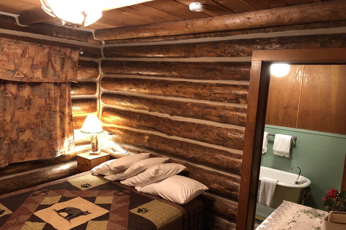 Habitación de cabaña de madera con cama, mesita de noche con lámpara, colcha, pared de madera y baño con lavamanos y bañera.