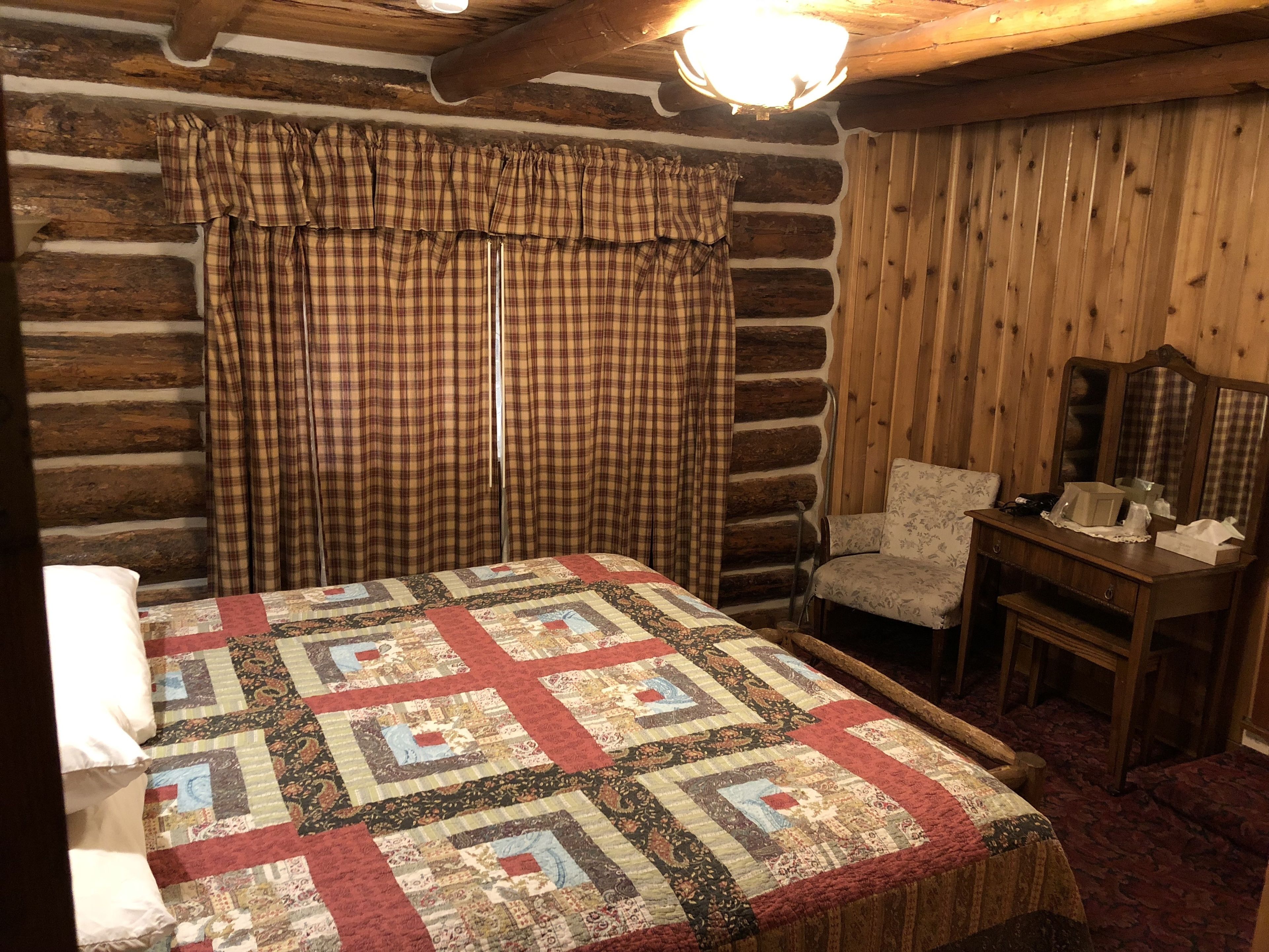 A log cabin bedroom with a quilted bedspread, plaid curtains, a wooden chair, and a nightstand with a lamp.
