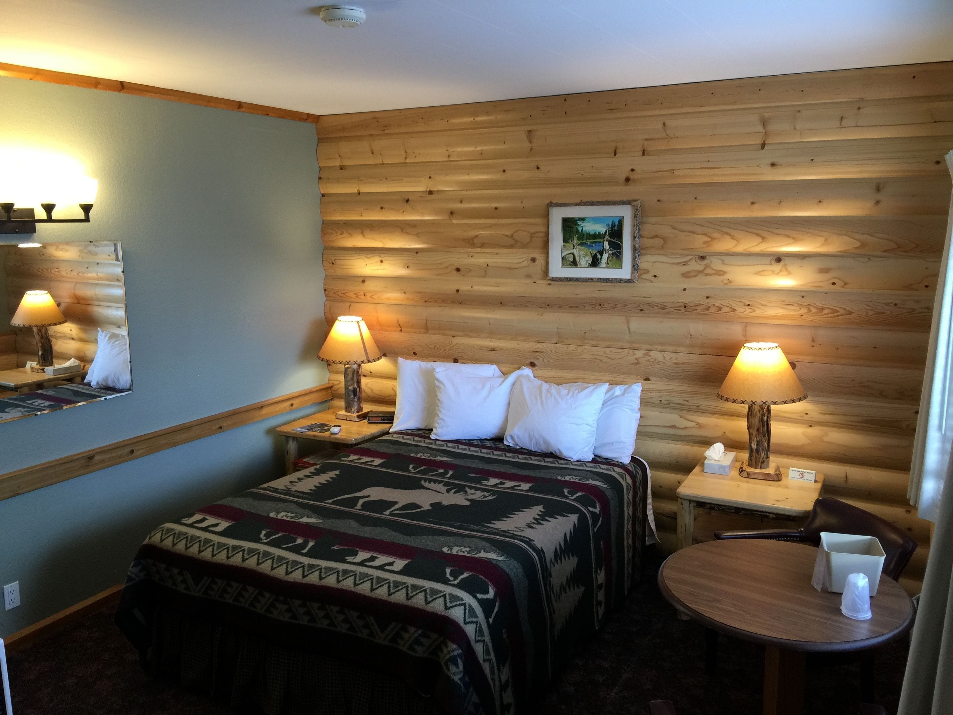 Una habitación acogedora con una pared de madera, una cama con una colcha estampada, dos lámparas de noche, una mesita con una taza y un cuadro en la pared.