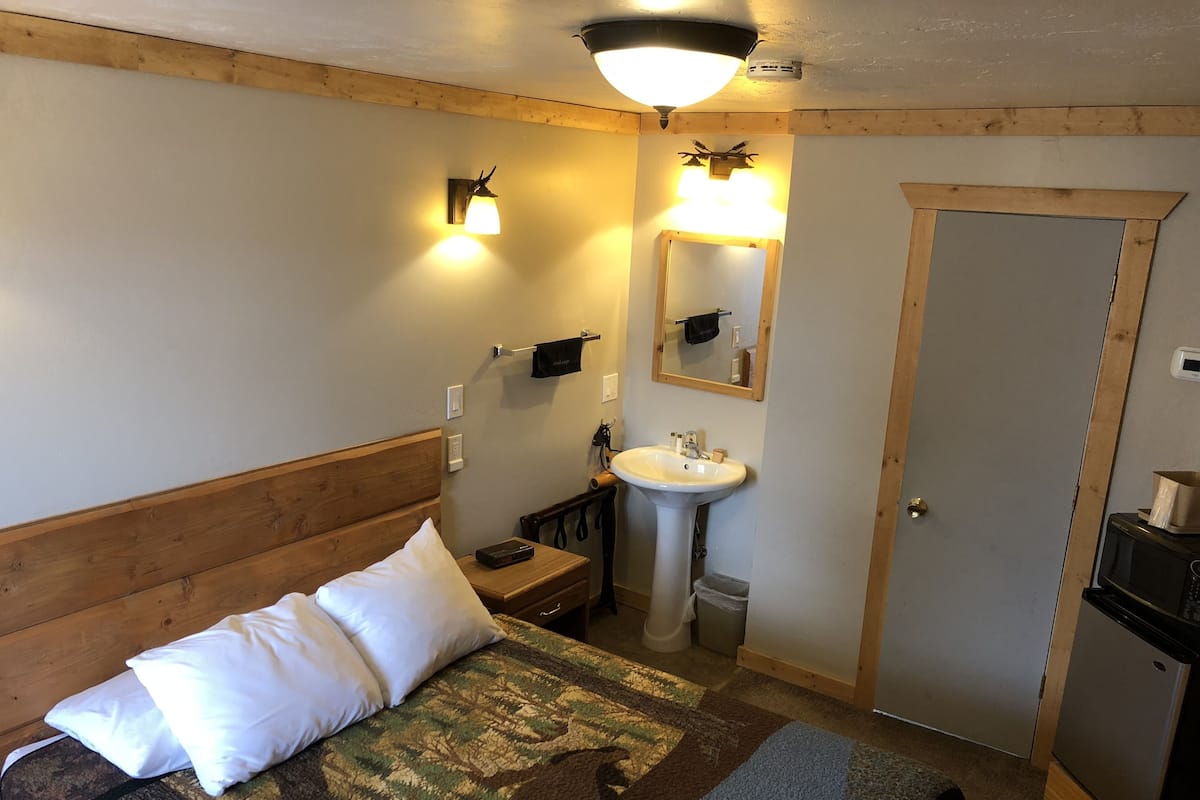 Un dormitorio con cabecera de madera, una cama con almohadas blancas, una mesita de noche con lámpara, un baño con lavamanos y espejo, y una puerta.