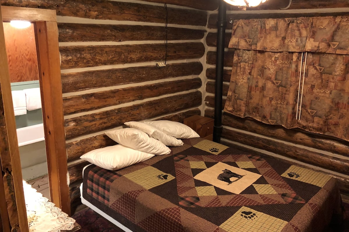 Un dormitorio de cabaña de troncos con una cama, almohadas, colcha, armario de madera y un espejo.