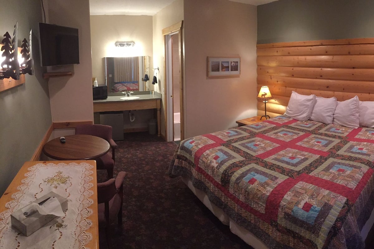 Habitación de hotel con cabecera de madera, cama con colcha de colores, mesita de noche con lámpara, escritorio con silla y televisor montado en la pared.