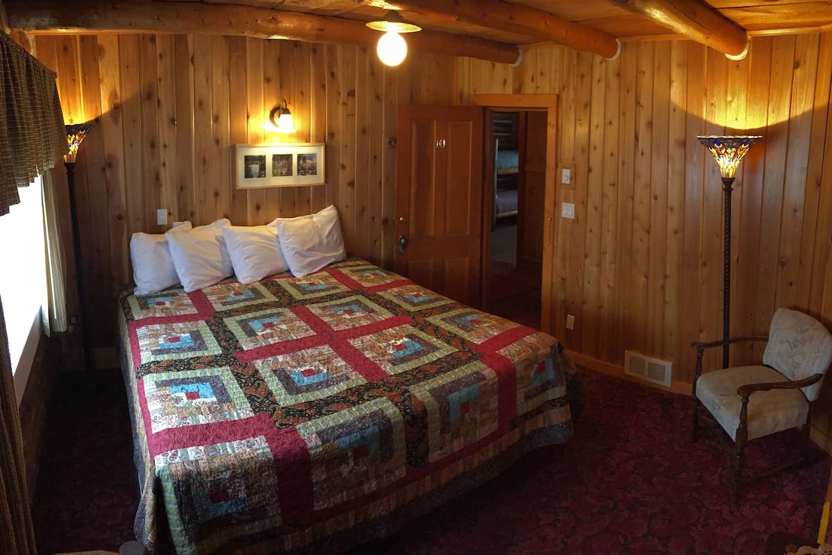 Una habitación con paneles de madera, una cama, un sillón y una lámpara fijada a la pared.