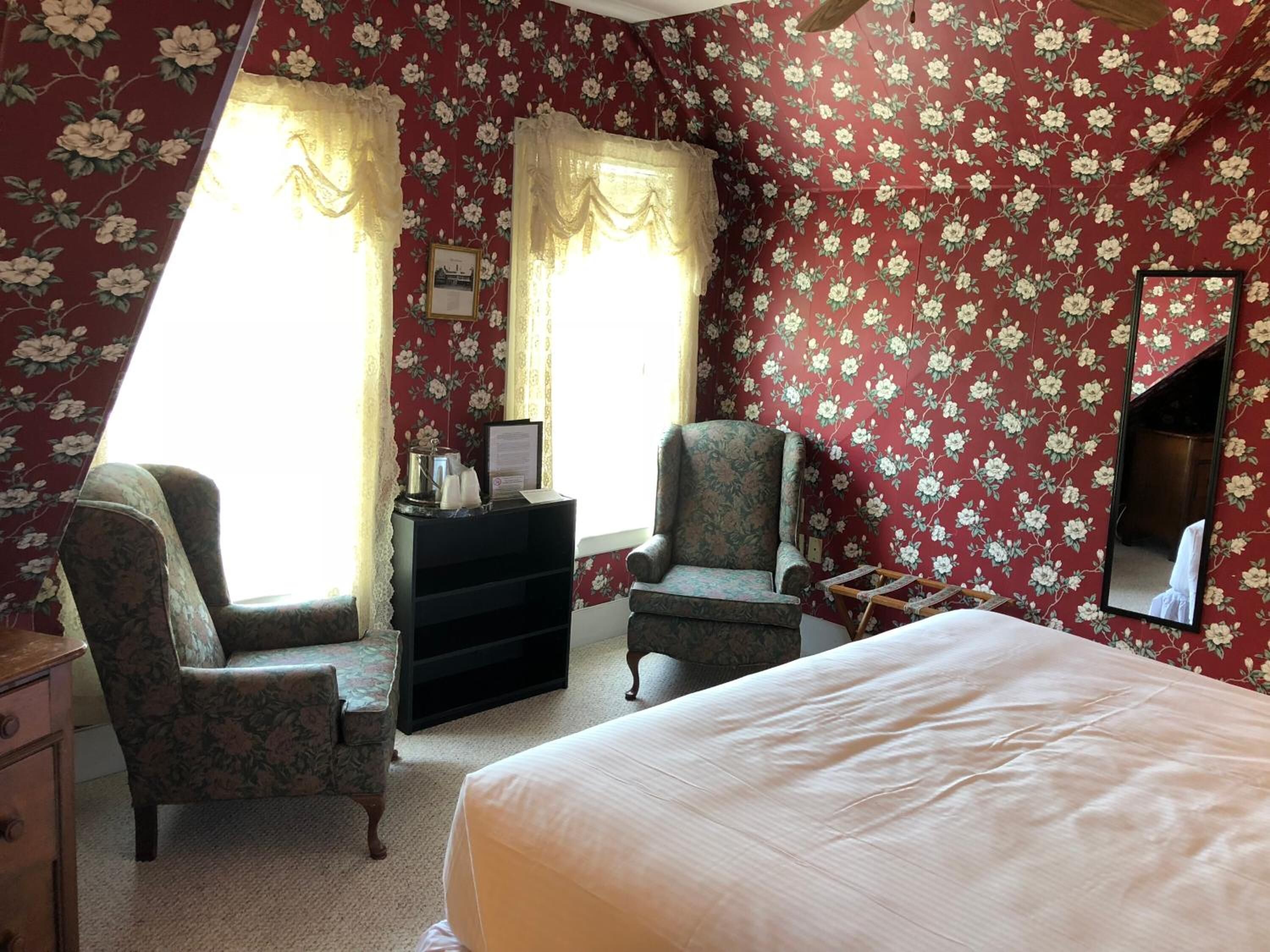 Een kamer met bloemenbehang, twee fauteuils, een bed, een dressoir en een spiegel.