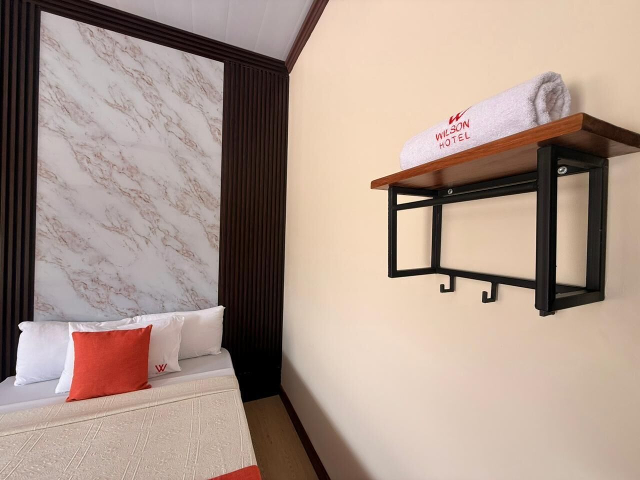 A hotel room with a bed, a marble-patterned wall, a wooden shelf with a towel, and a black metal shelf.
