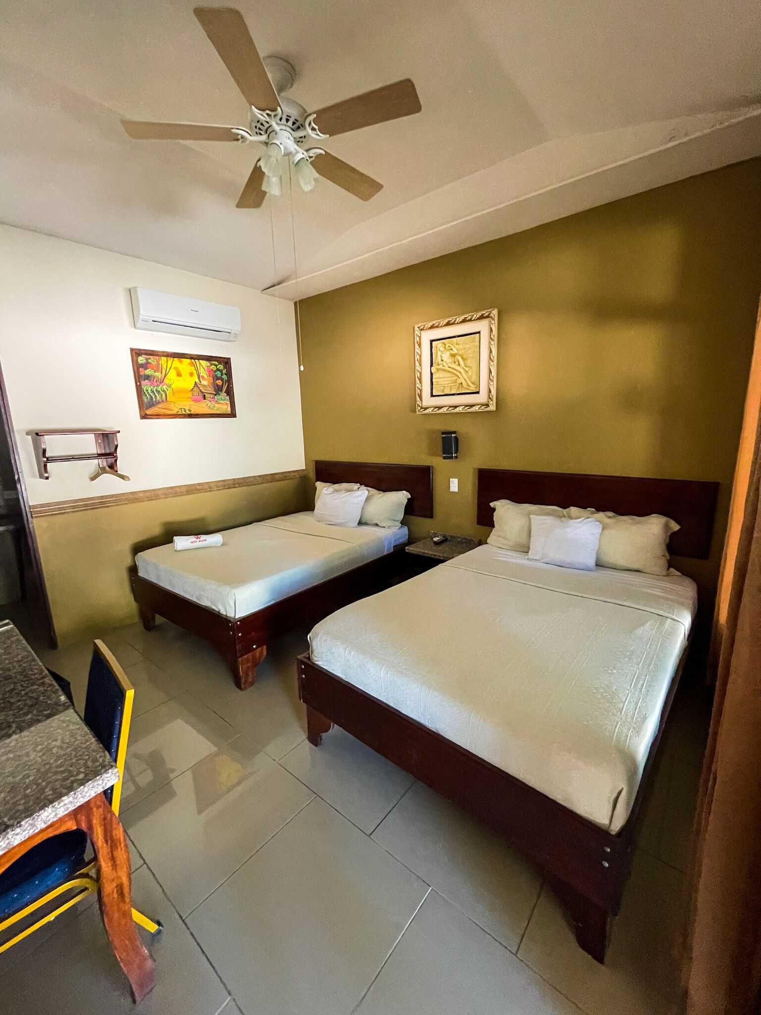 Une chambre d’hôtel avec deux lits, un ventilateur de plafond, un climatiseur, une petite table et une chaise.