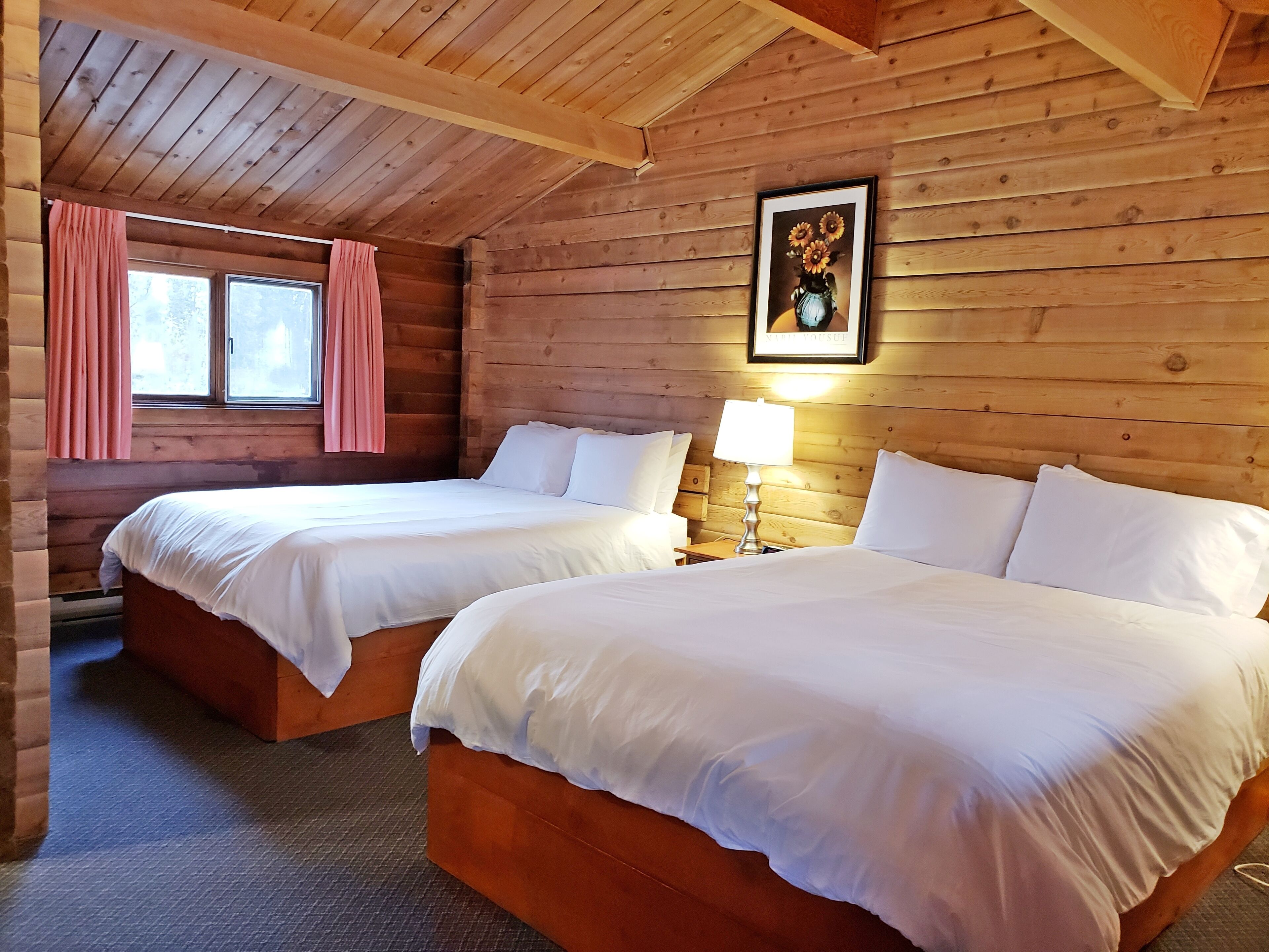 A log cabin room with two beds, a window with pink curtains, a framed picture on the wall, and a nightstand with a lamp.