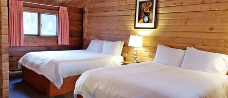 A log cabin room with two beds, a window with pink curtains, a framed picture on the wall, and a nightstand with a lamp.
