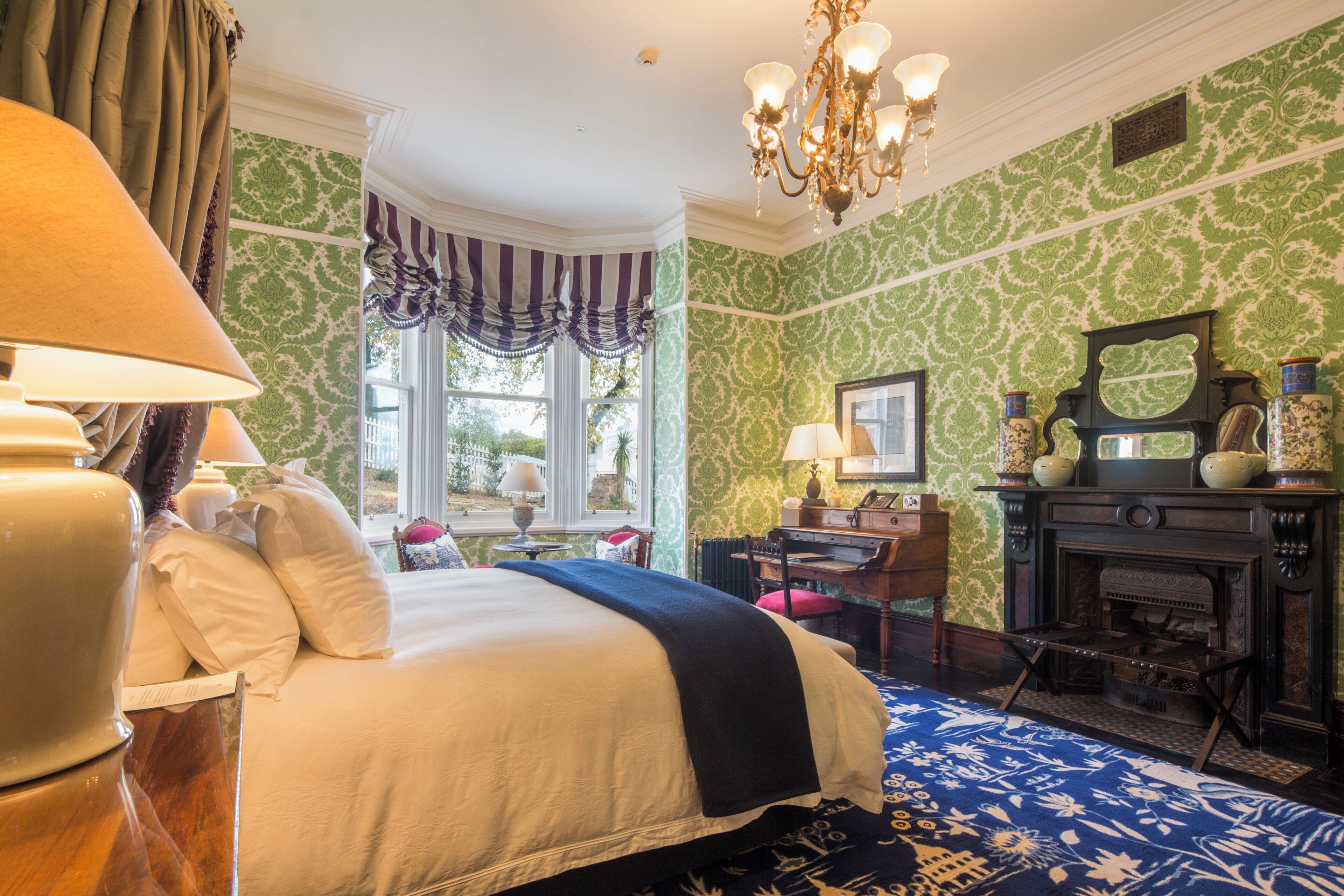 A room with a large bed, a fireplace, a chandelier, and green patterned wallpaper.