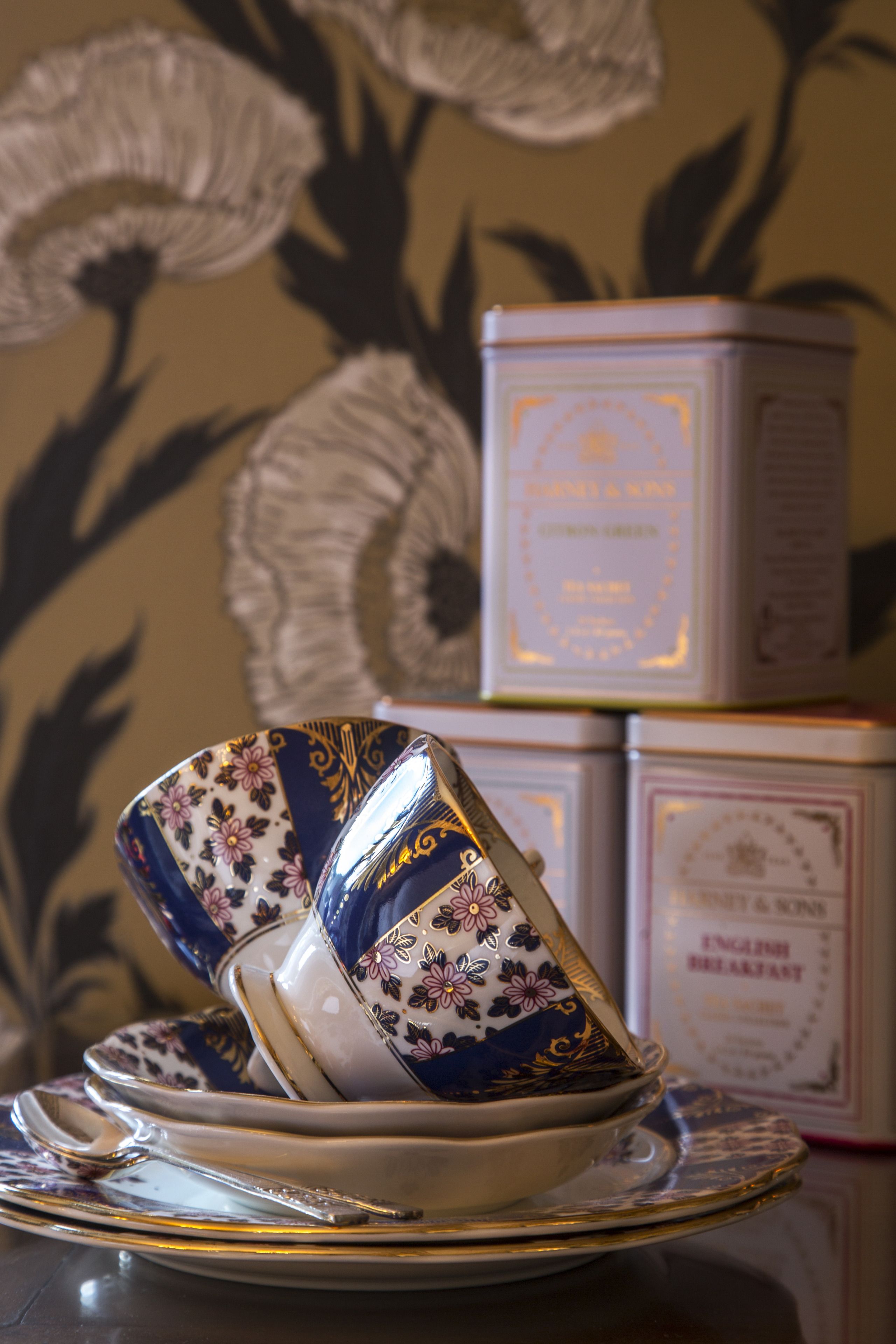 Una pila de cajas de té con diseños florales, una taza y platillo, y una cuchara sobre una superficie reflectante.