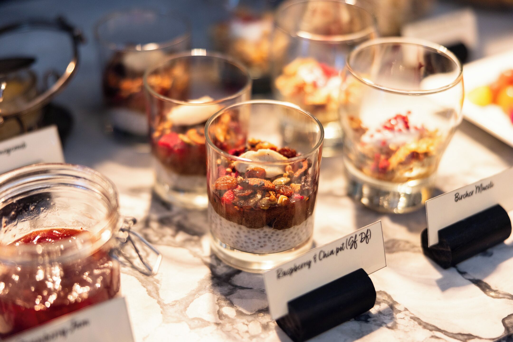 Diversos doces em copos transparentes com etiquetas, incluindo 'Raspberry & Chia pudding' e 'Burchard Muesli'.