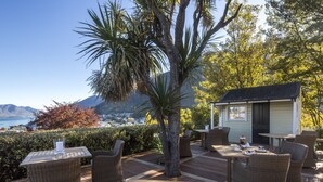 An outdoor patio with wicker furniture, a small building, and a view of the sea and mountains. - Hulbert House (Queenstown)