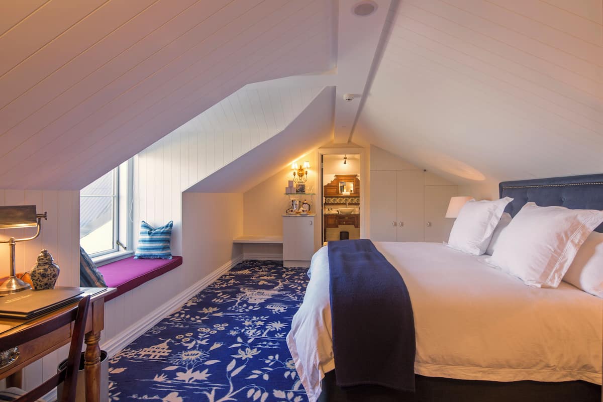 Un dormitorio con techo inclinado, cama con ropa blanca, asiento de ventana con cojín azul y escritorio con lámpara.
