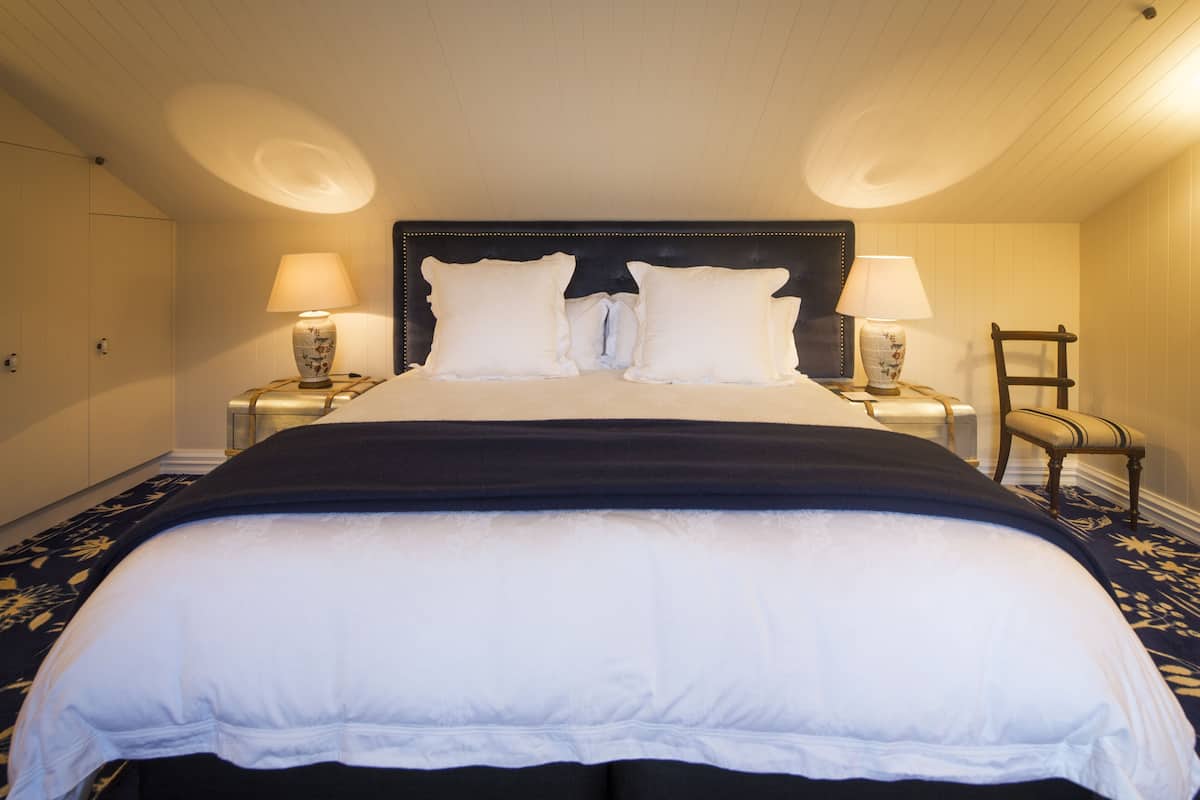 Una cama bien tendida con un cabecero oscuro y ropa de cama blanca, flanqueada por dos mesitas de noche con lámparas.
