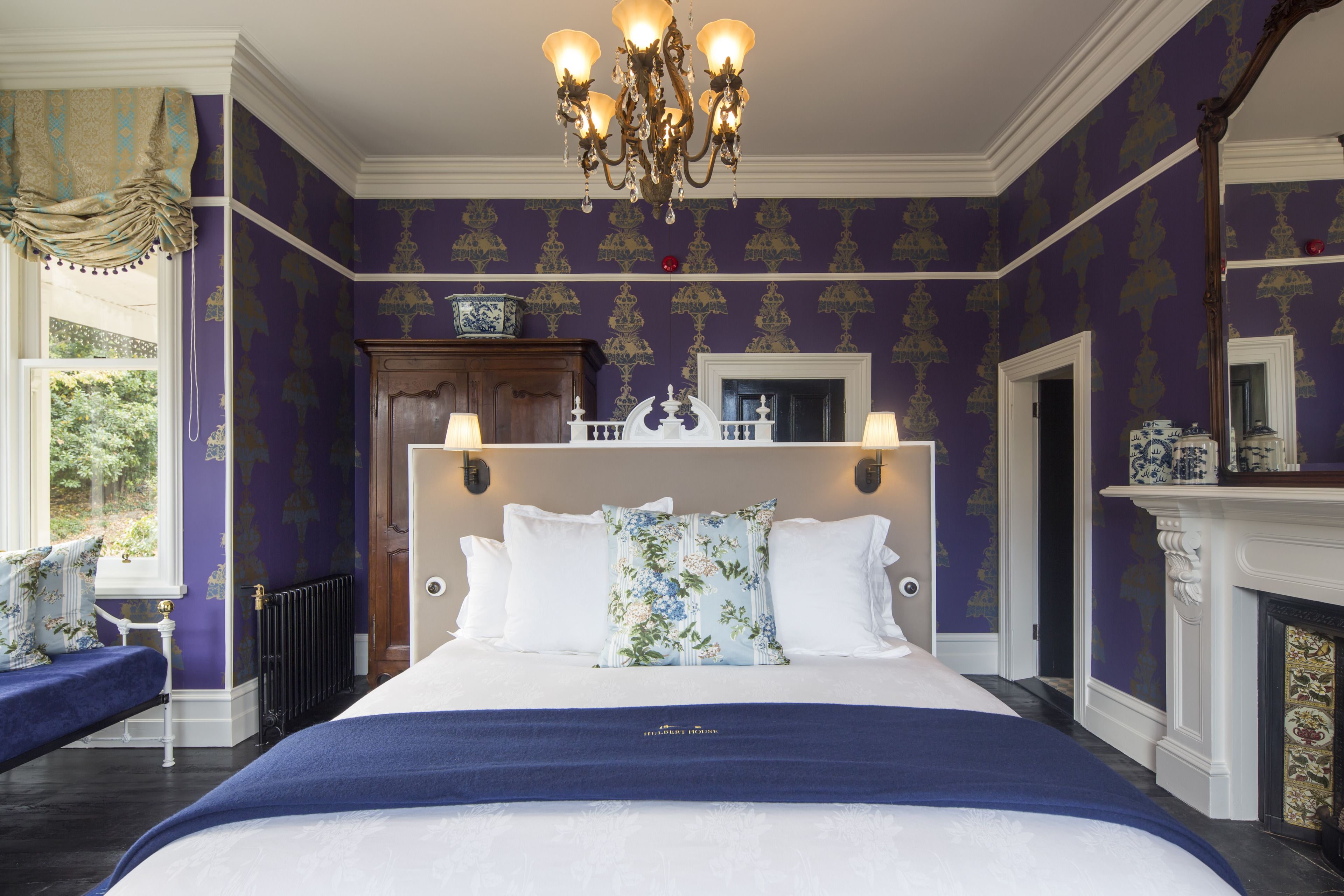 A large bed with white linens, a floral pillow, and a blue throw blanket. There is a chandelier above the bed and a fireplace to the right.