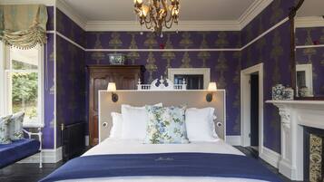 A large bed with white linens, a floral pillow, and a blue throw blanket. There is a chandelier above the bed and a fireplace to the right.