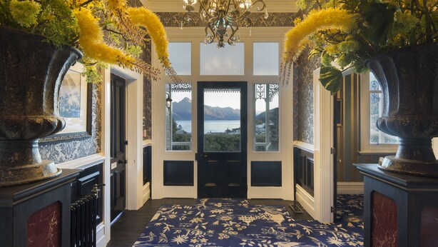 A hallway with a view of mountains through large windows, a chandelier, and floral arrangements on either side. - Hulbert House (Queenstown)
