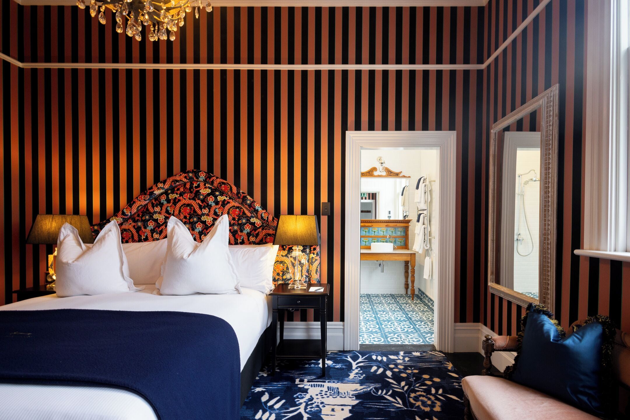 Een hotelkamer met gestreepte behang, een bed met wit en blauw beddengoed, een bed met een gedessineerd hoofdbord, een nachtkastje met een lamp en uitzicht op de badkamer.