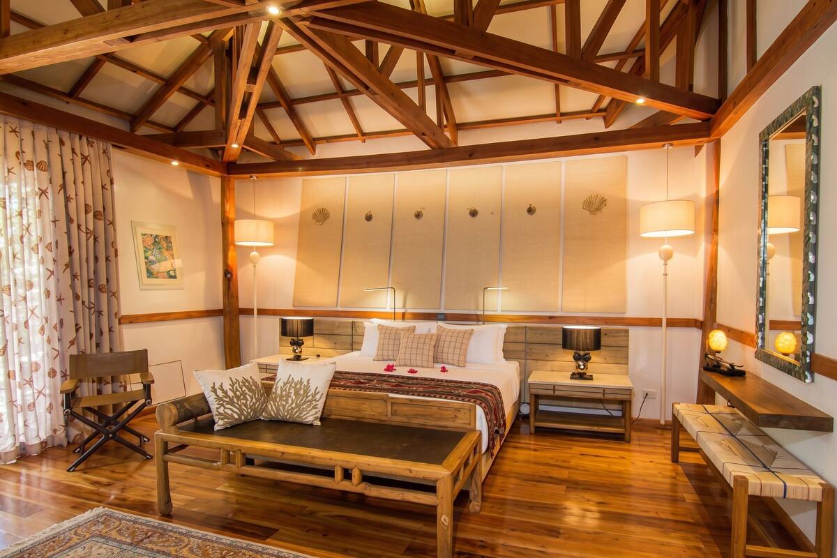 A room with a wooden ceiling, a bed with a headboard, bedside tables, a bench, and a patterned rug.