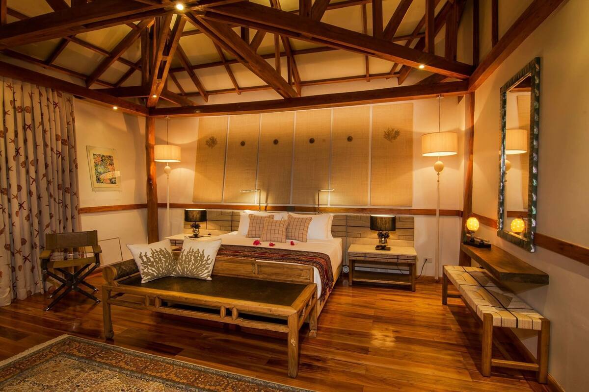 A bedroom with a wooden ceiling, a bed with multiple pillows, bedside tables with lamps, a bench, and a patterned rug.