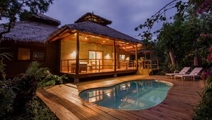 Una casa de madera con techo de paja, piscina y terraza cubierta.
