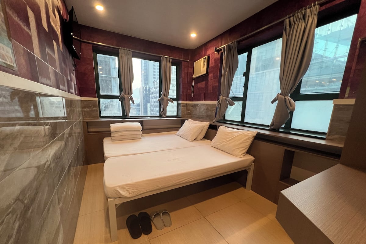 A hotel room with two beds, a window with curtains, a pair of slippers, and a view of buildings outside.
