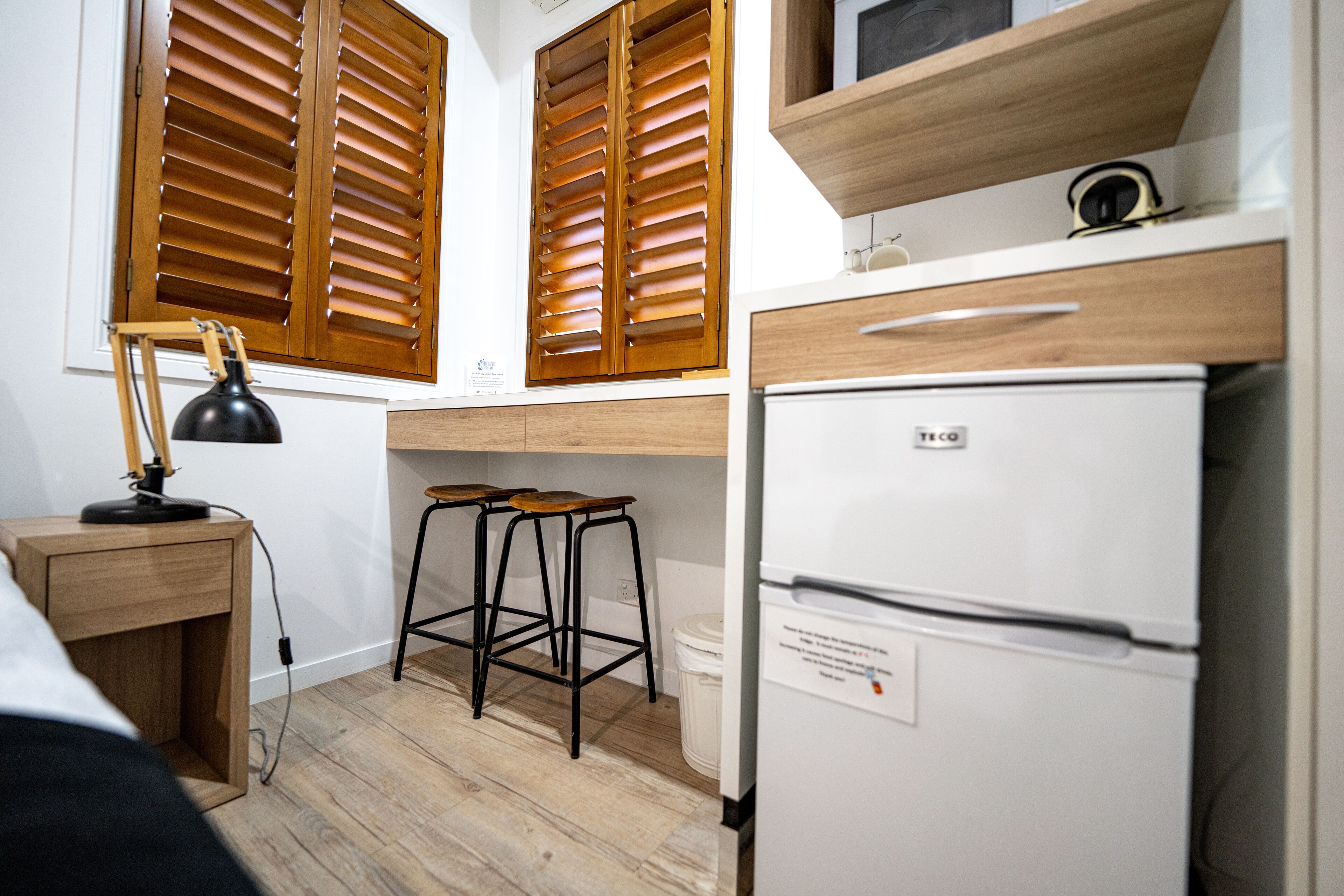 Une cuisine de taille réduite avec des volets en bois, un réfrigérateur blanc, un tabouret de bar et un bureau avec une lampe.