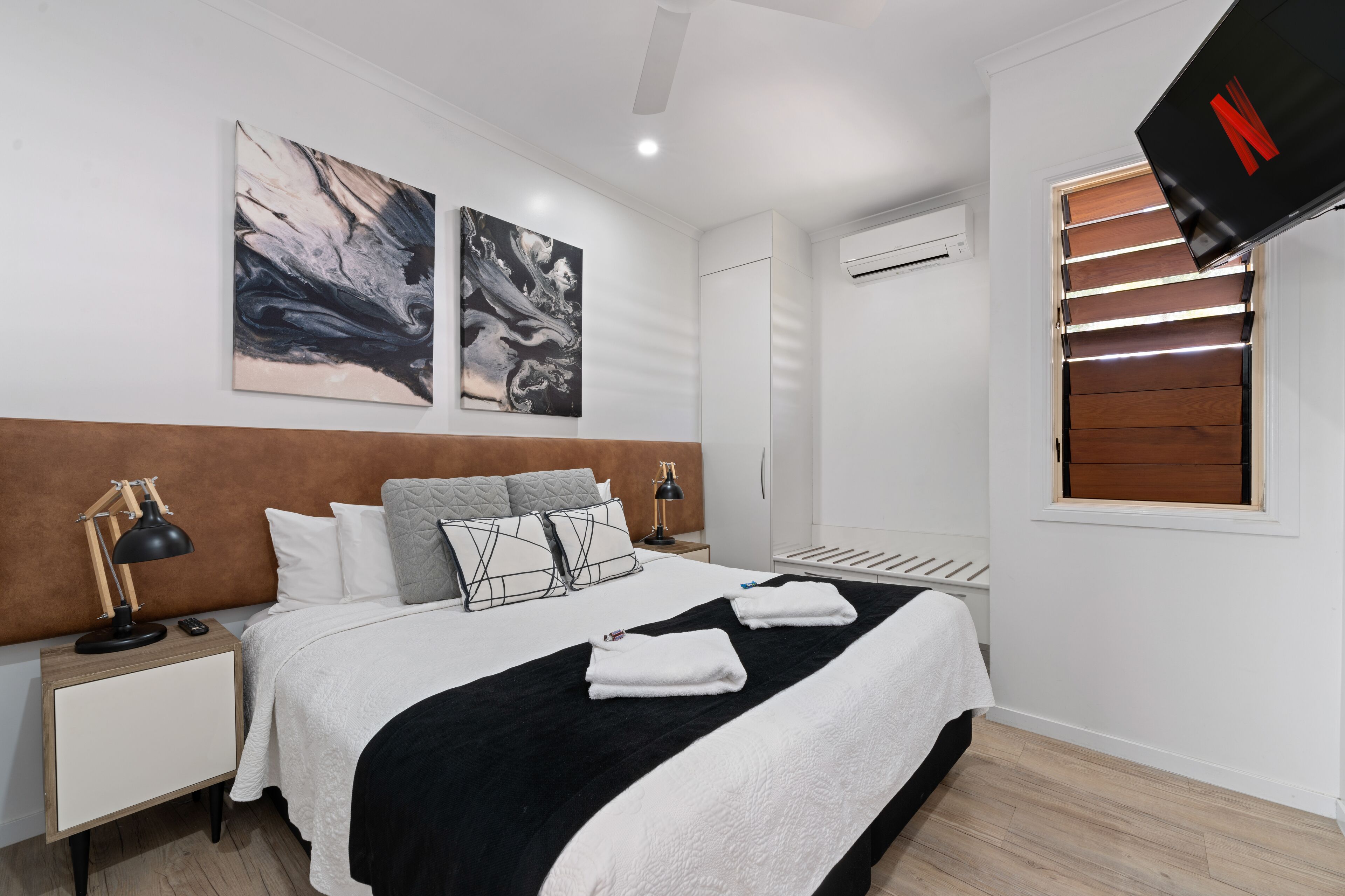 A modern hotel room with a large bed, two wall-mounted artworks, a ceiling fan, and a TV mounted on the wall.
